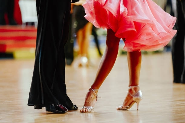 Dancers on a wooden floor, woman in pink dress and heels, man in black pants, ballroom setting.