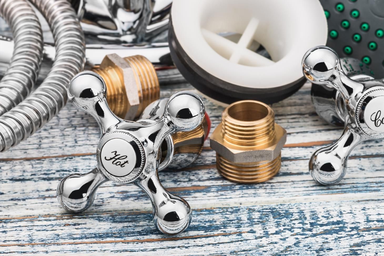 Close-up of plumbing fixtures: chrome faucet handles, brass fittings, drain, and shower hose on a weathered wooden surface.