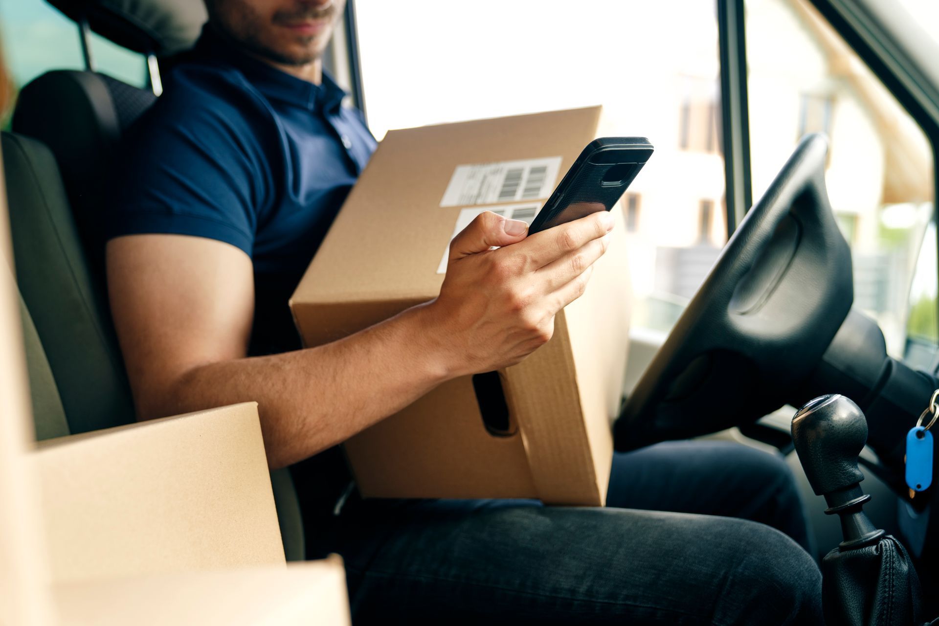 A delivery man is sitting in a van holding a cardboard box and using a cell phone.