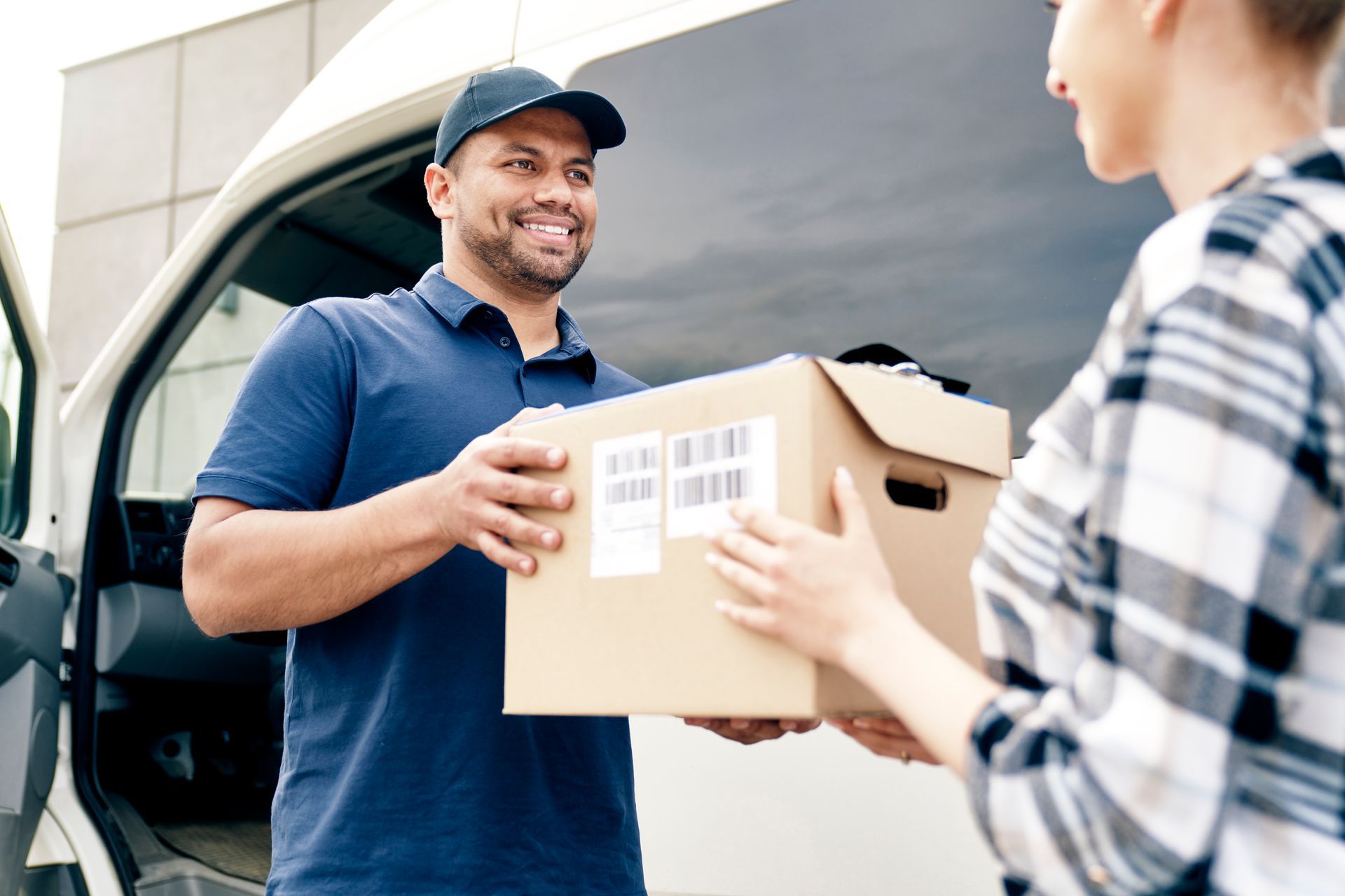 Delivery person handing a cardboard box to a person, beside a white van; both smiling.