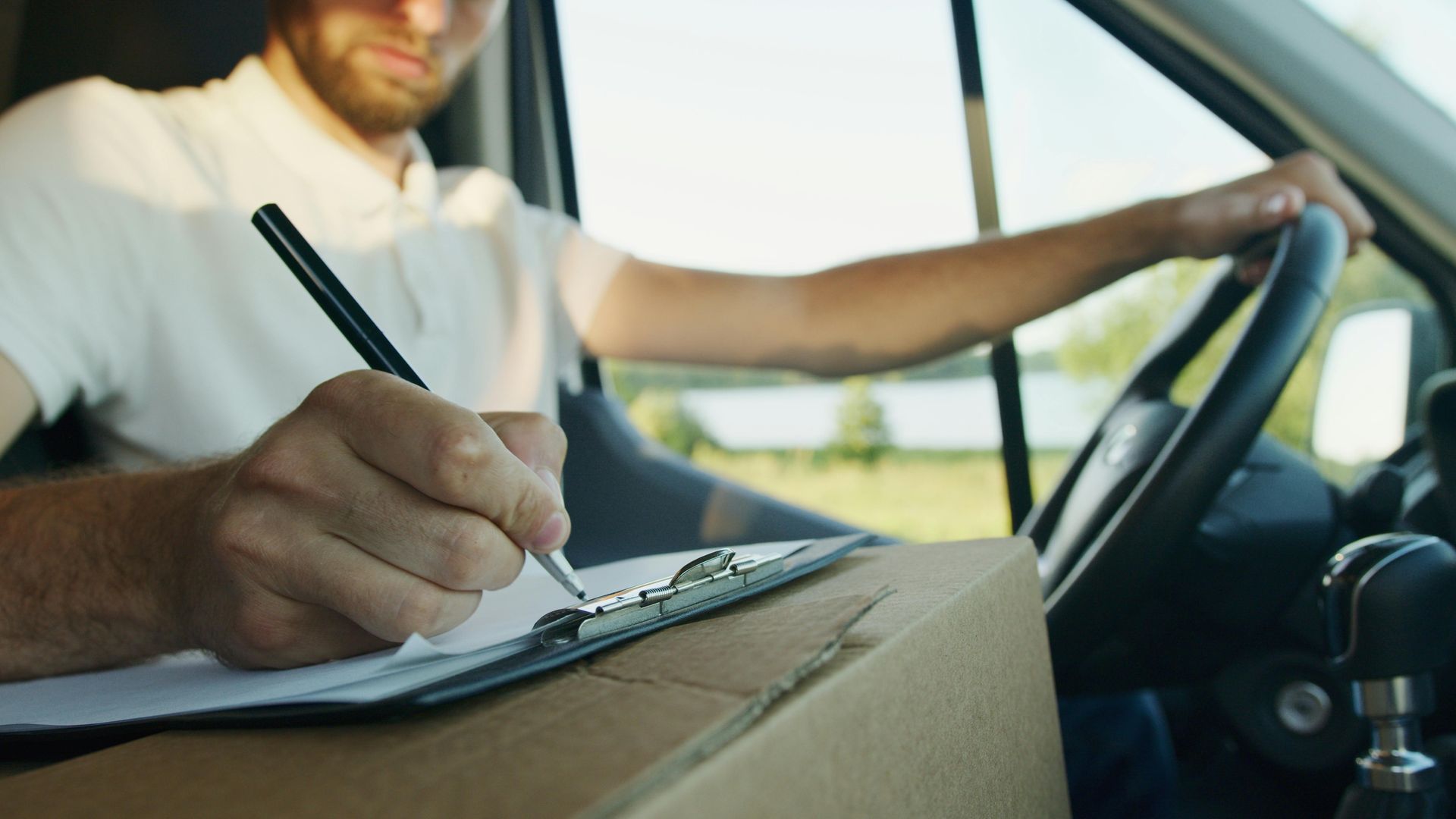 A delivery man is writing on a clipboard while driving a van.