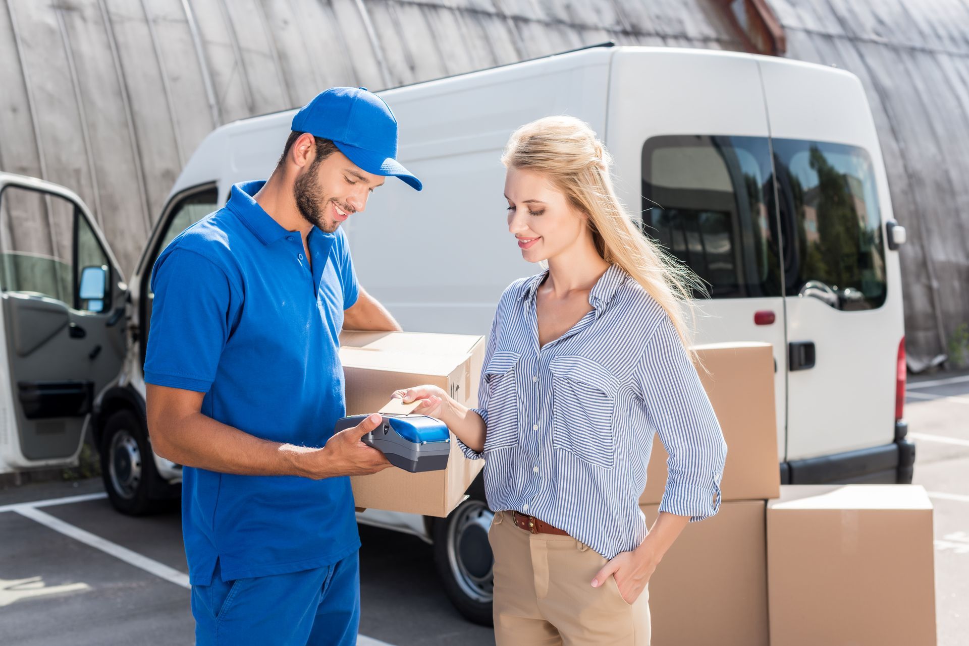 Delivery person in blue uniform hands payment terminal to a woman in a striped shirt, boxes on ground.