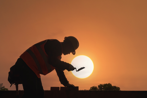 Construction worker laying bricks at sunset, wearing safety vest.