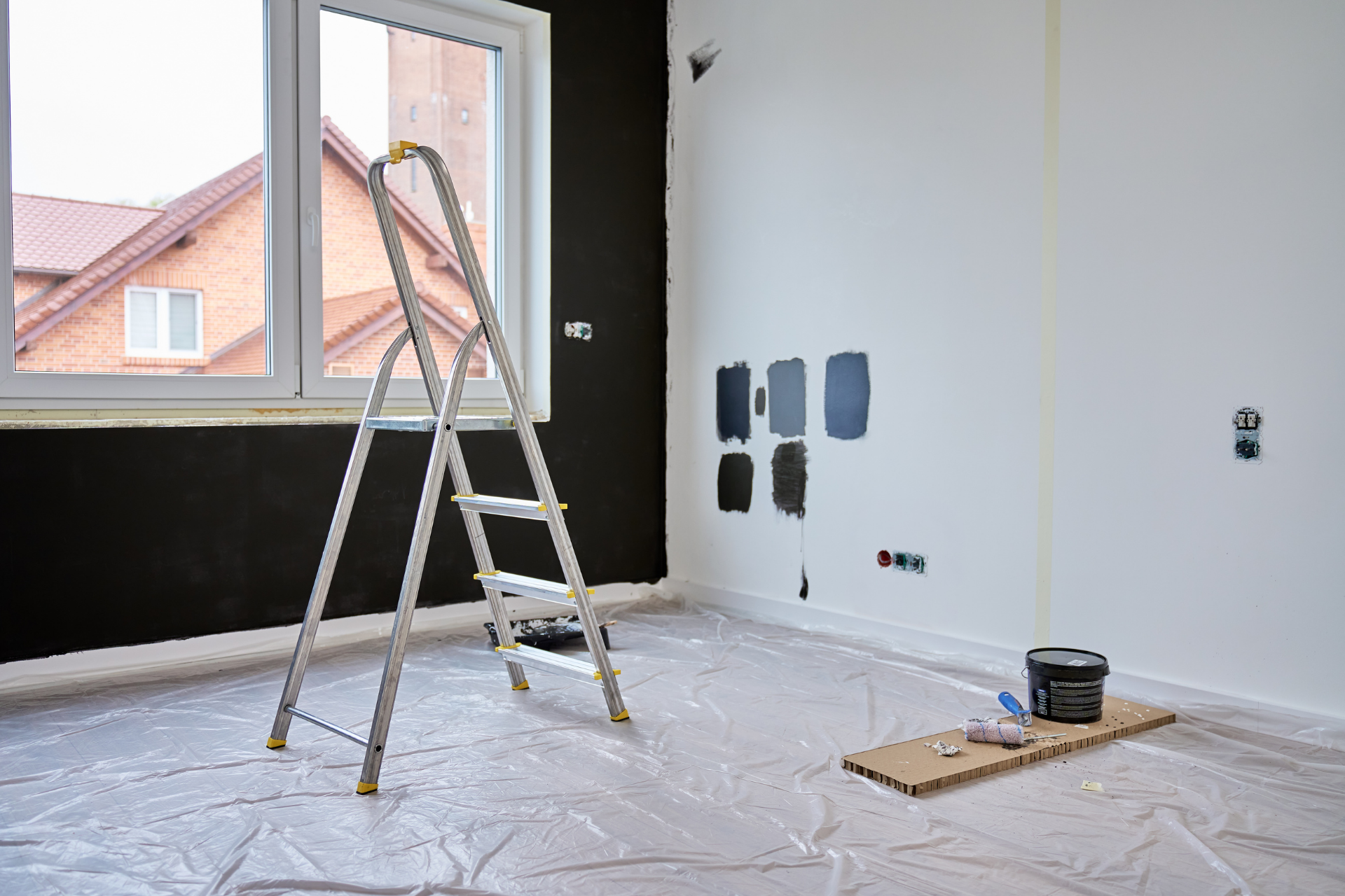 Room being painted: ladder, partially painted black wall, white walls with paint swatches, plastic sheeting on floor.