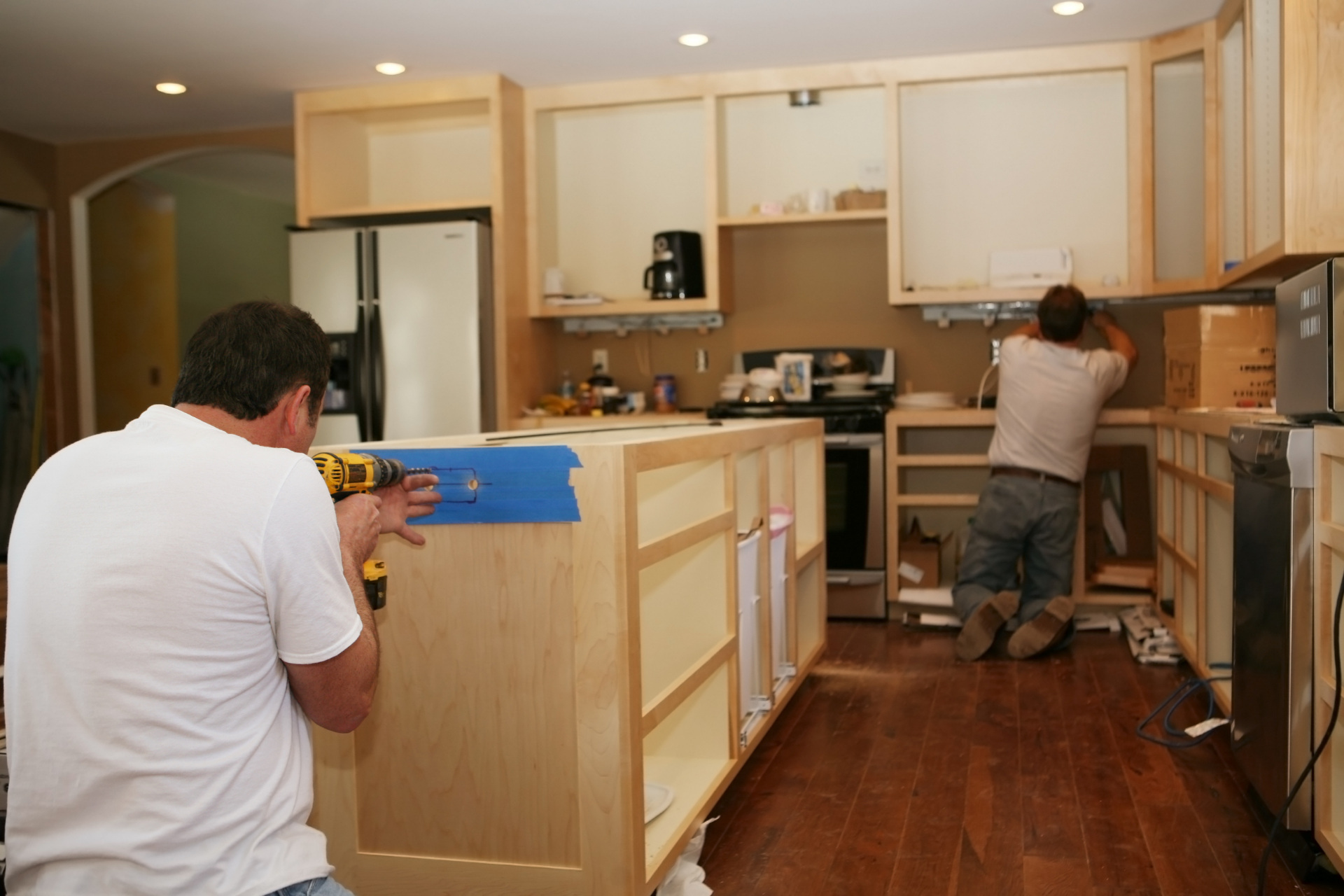 Two men installing light wood kitchen cabinets in a room with hardwood floors.
