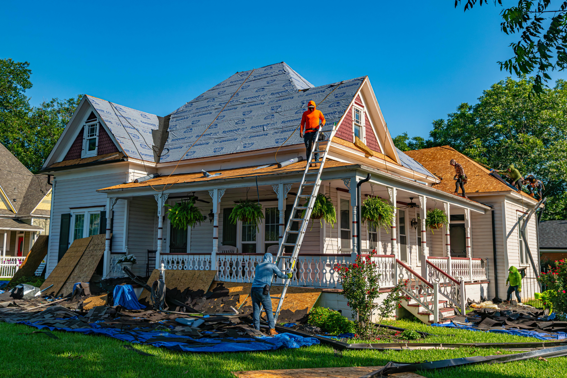 Roofers repairing a two-story white house with a porch. Workers on ladders and roof installing shingles on a sunny day.