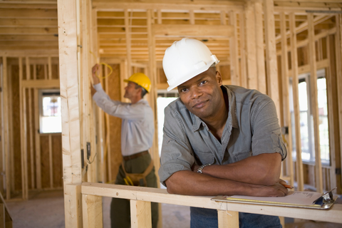 Construction worker leaning on railing, smiling; another worker in background. Interior, wooden framework.