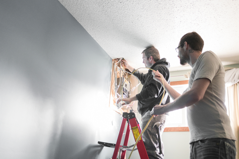 Two men working on electrical wiring near a ceiling, one on a ladder, in a room with blue walls.