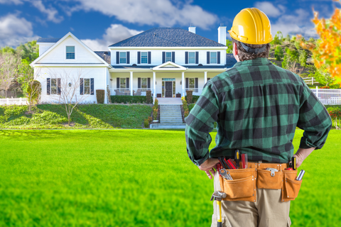 Construction worker in hardhat, facing large white house, hands on hips.