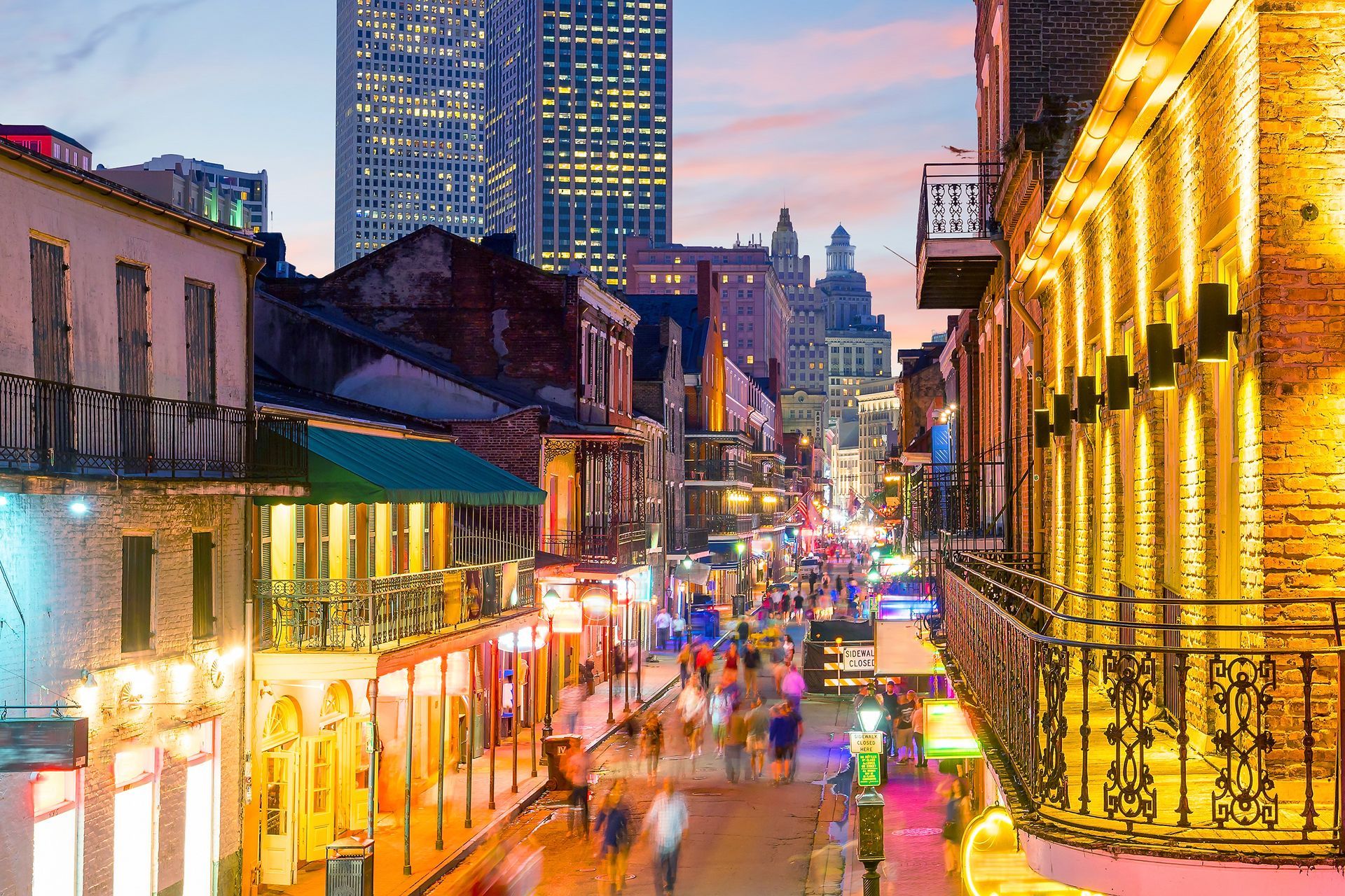 Colorful neon-lit city street at dusk with crowded sidewalks and historic buildings in the background