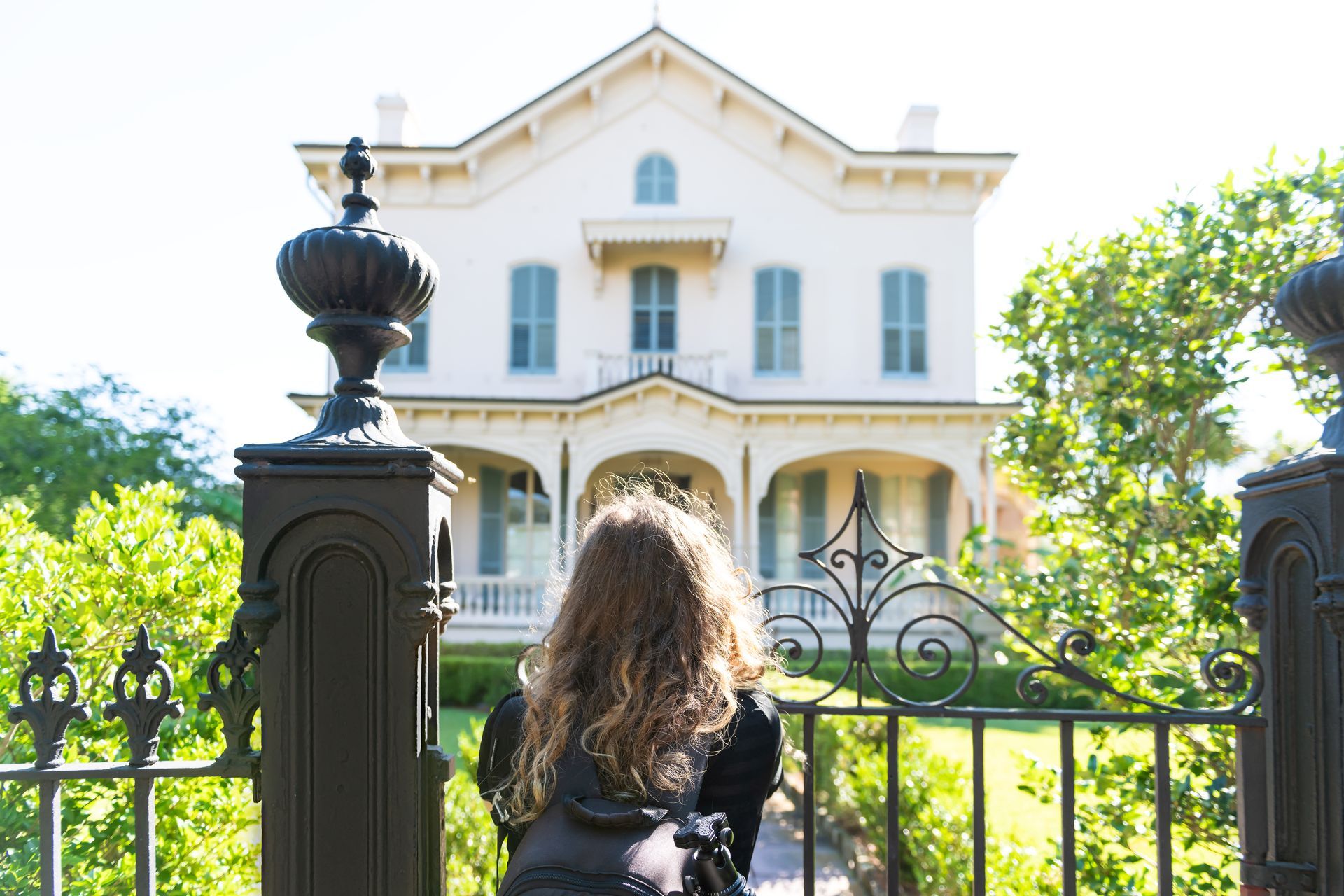 Person facing a white house behind a black gate, with bright green trees in front.