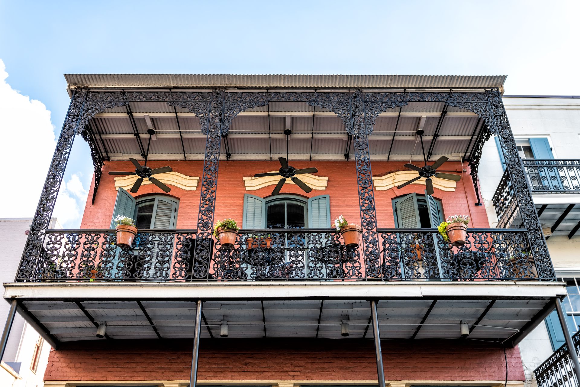 Balcony of a brick building with ornate iron railings, ceiling fans, and potted greenery
