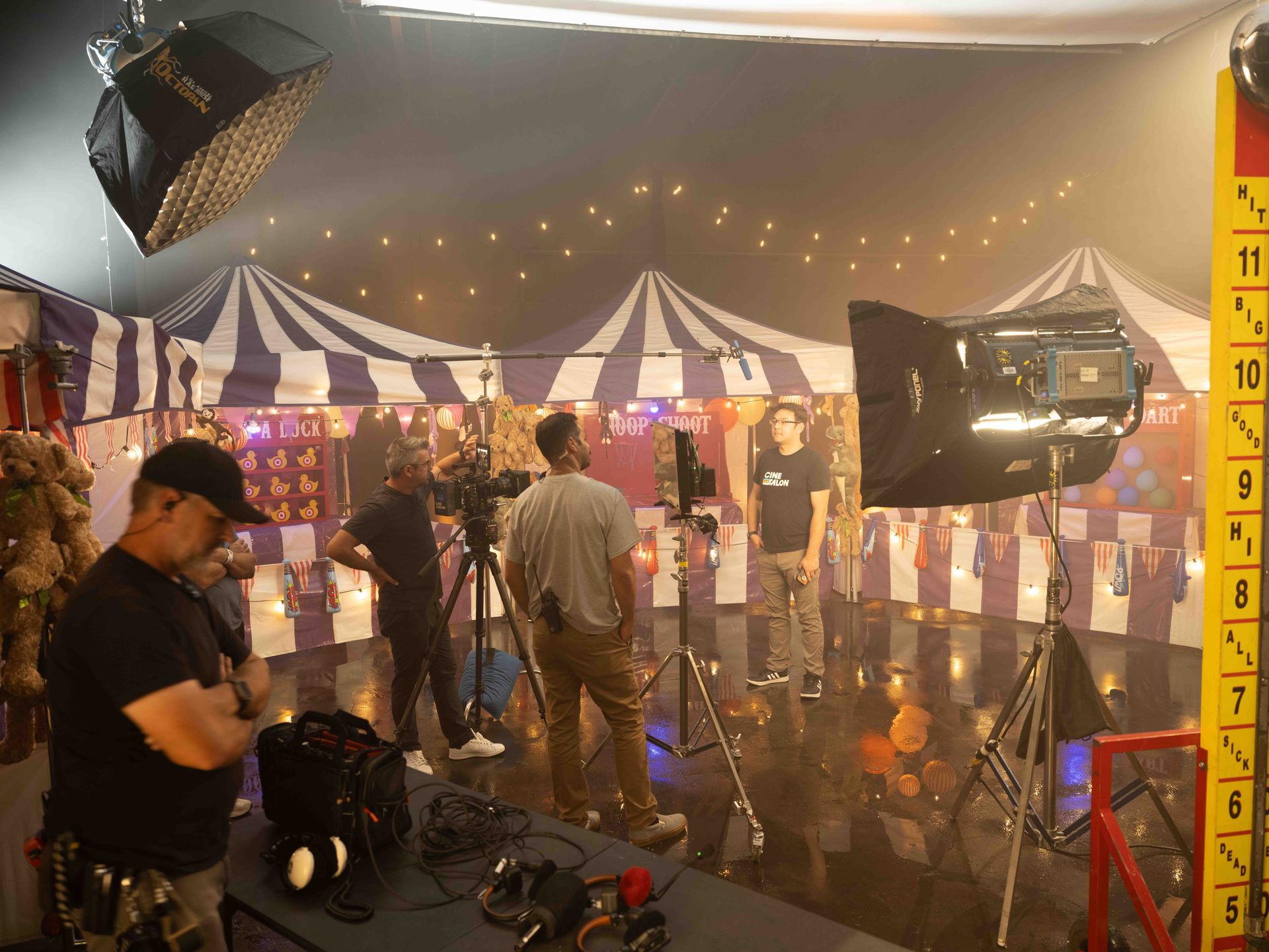 Film set: Crew filming person in front of a carnival-themed backdrop. Lighting equipment visible.