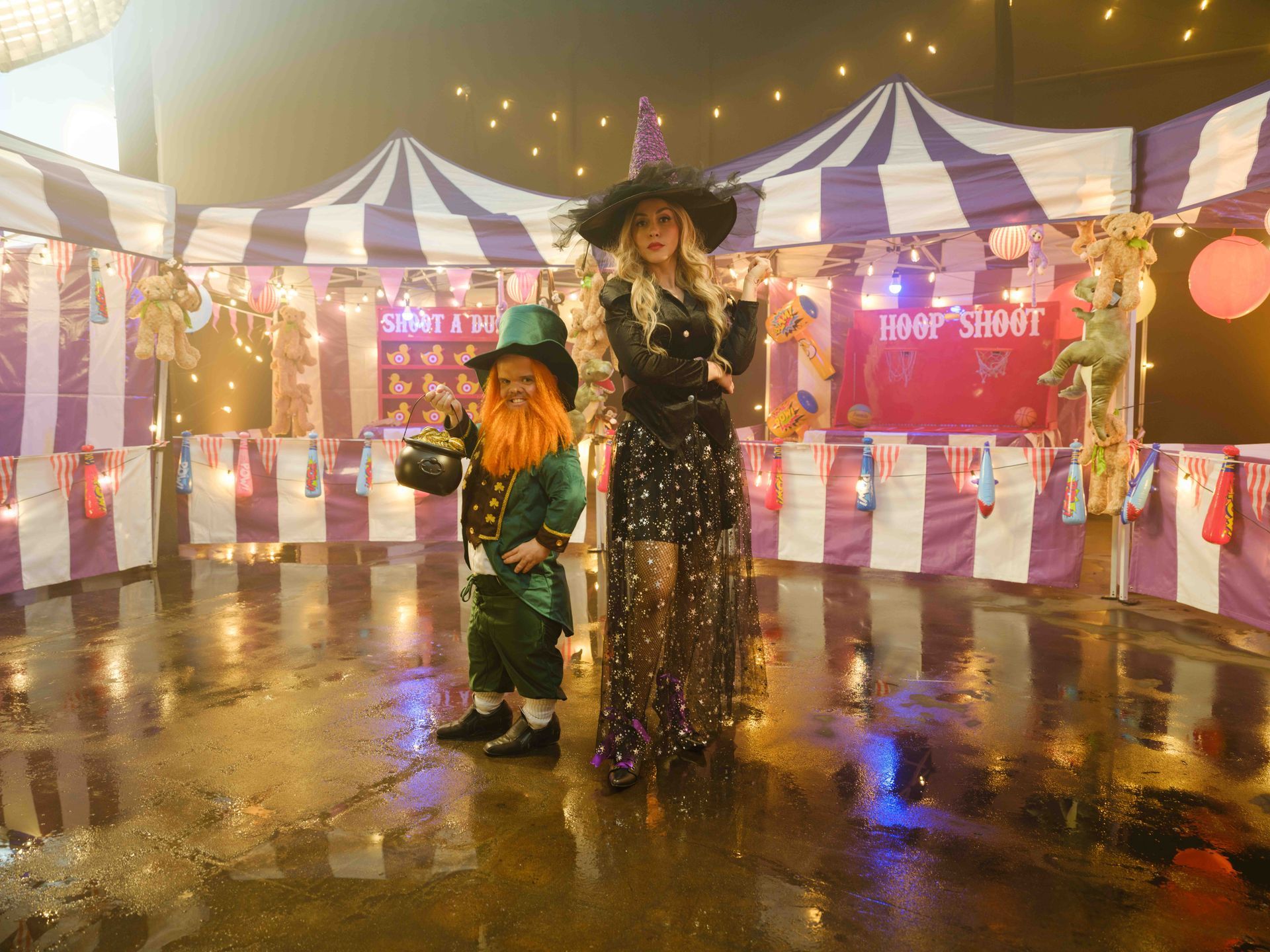 Woman in witch costume and leprechaun stand in front of carnival game booths.