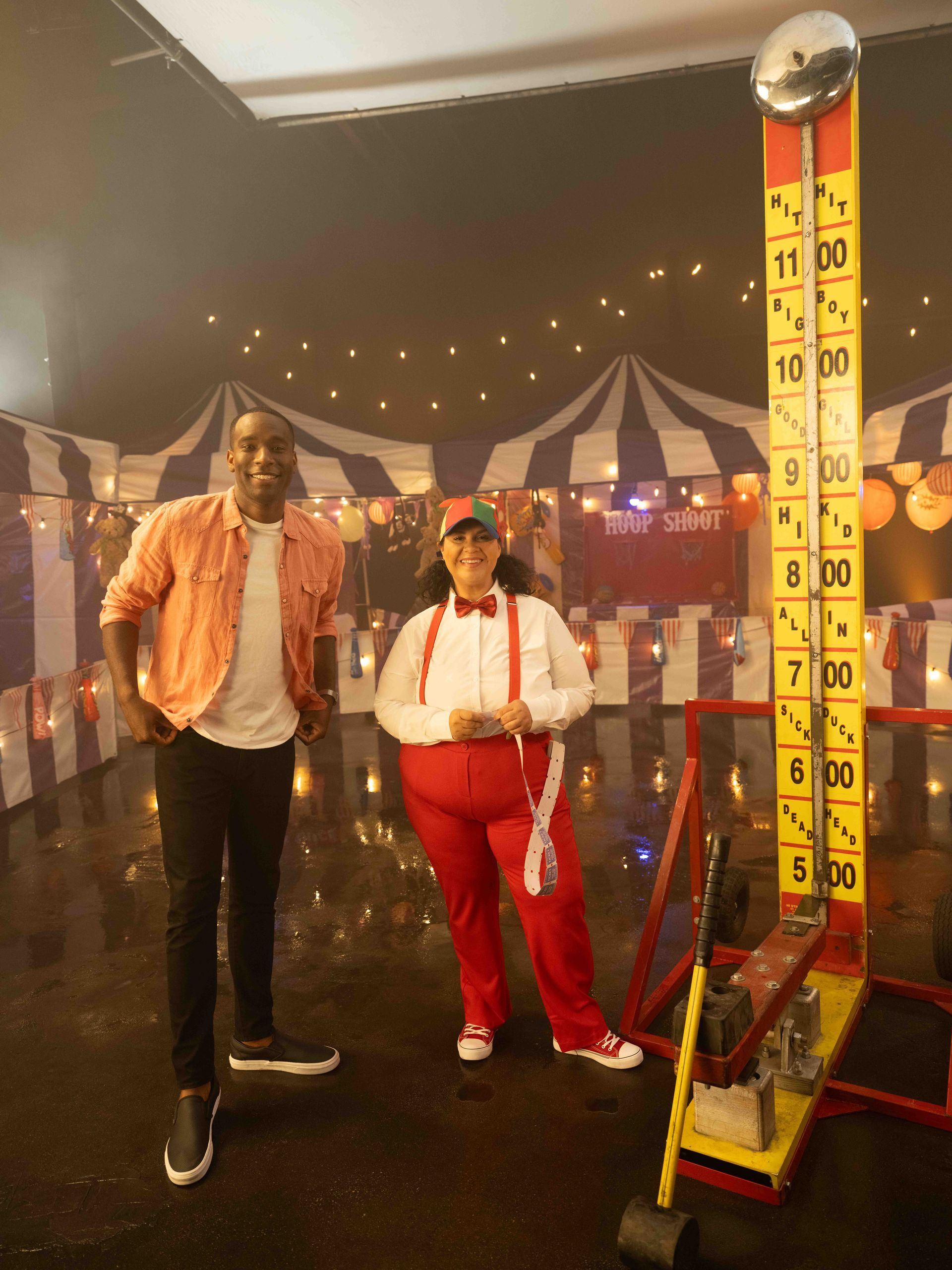 Two people pose next to a high striker carnival game. Man wears a shirt and pants, woman is in a red outfit.