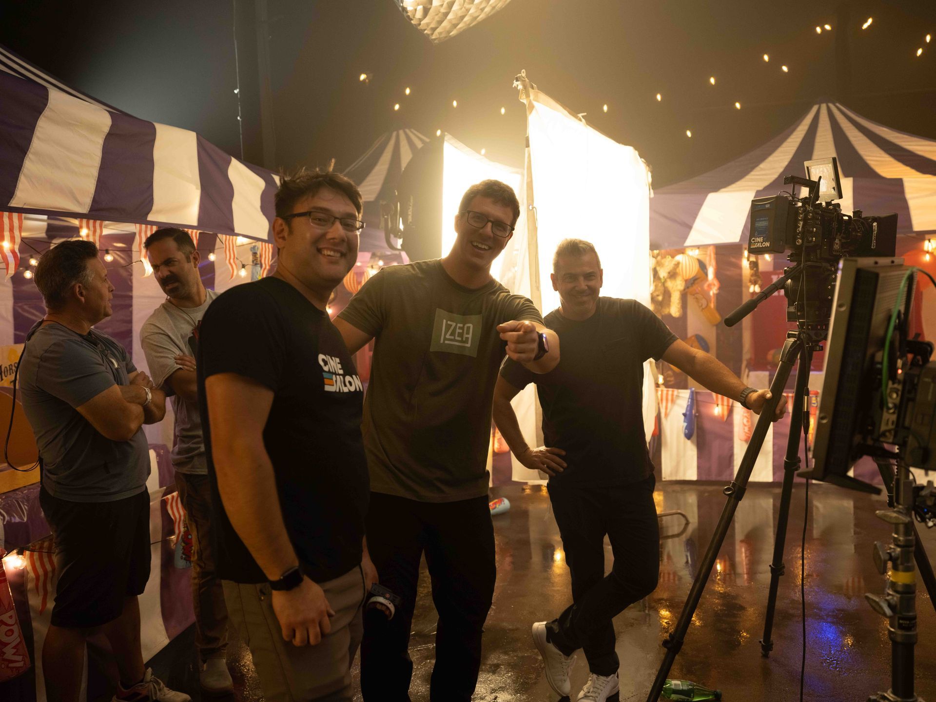 Film crew on set, laughing, pointing. Circus tents in background, bright lights, camera on tripod.