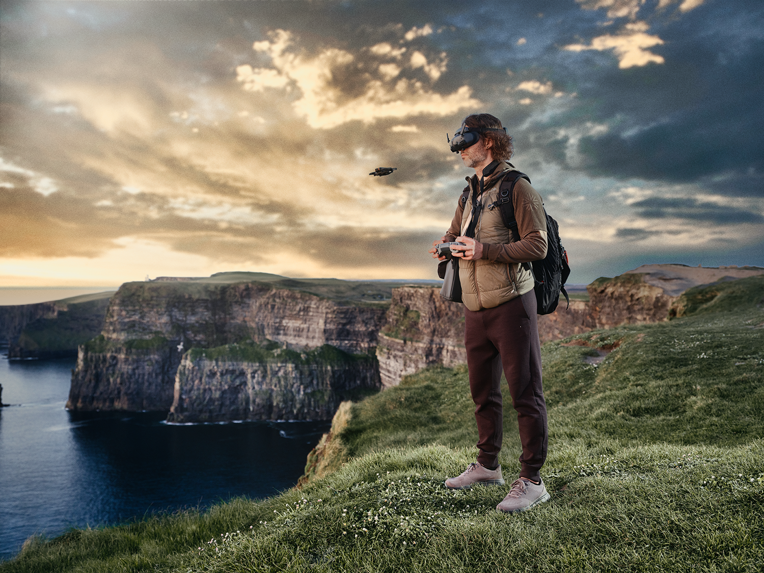 Person wearing VR headset stands on a cliff overlooking ocean. Drone hovers in sky. Sunset.