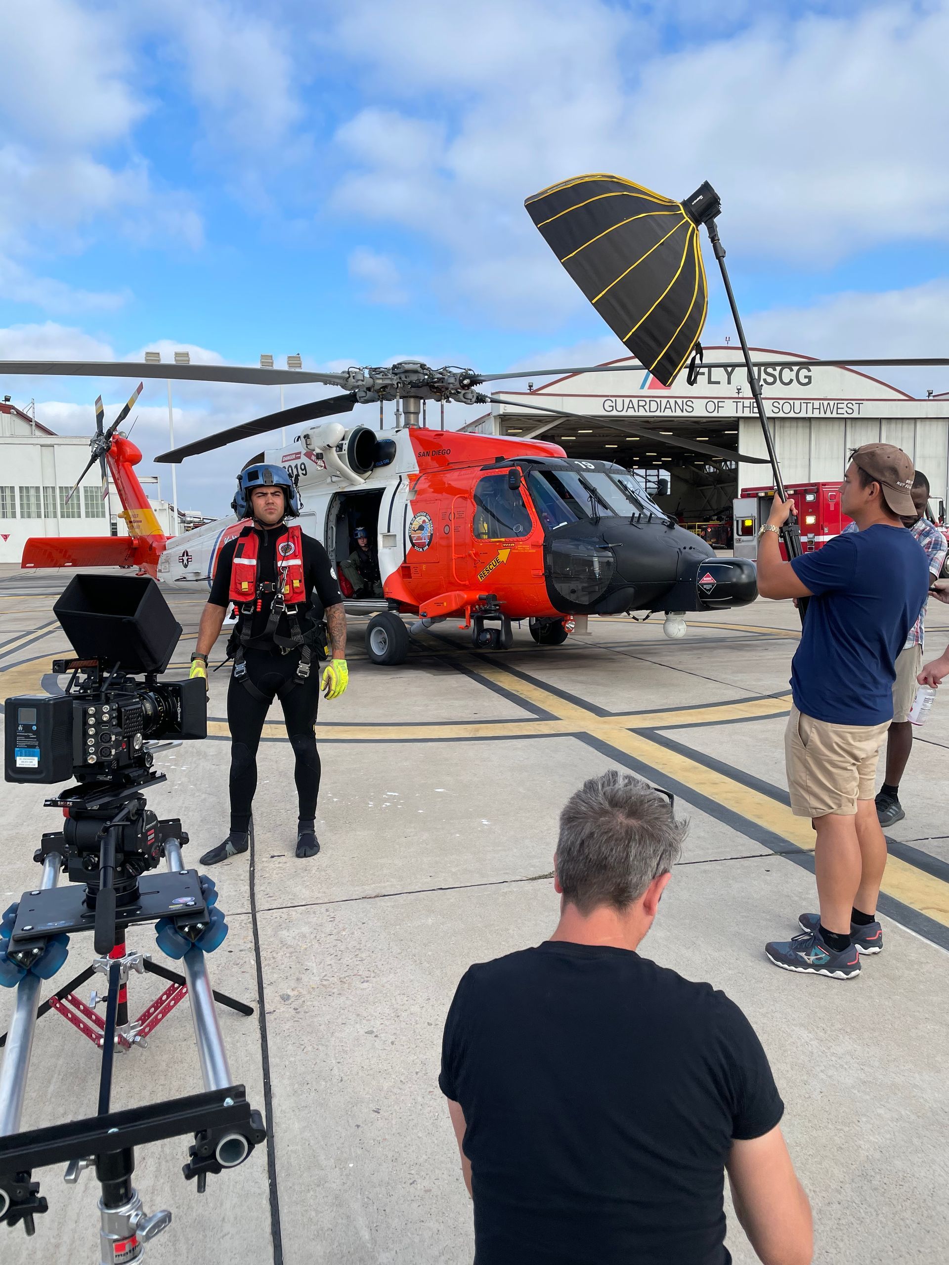Coast Guard helicopter on tarmac; crew member stands ready as filming occurs, holding equipment.