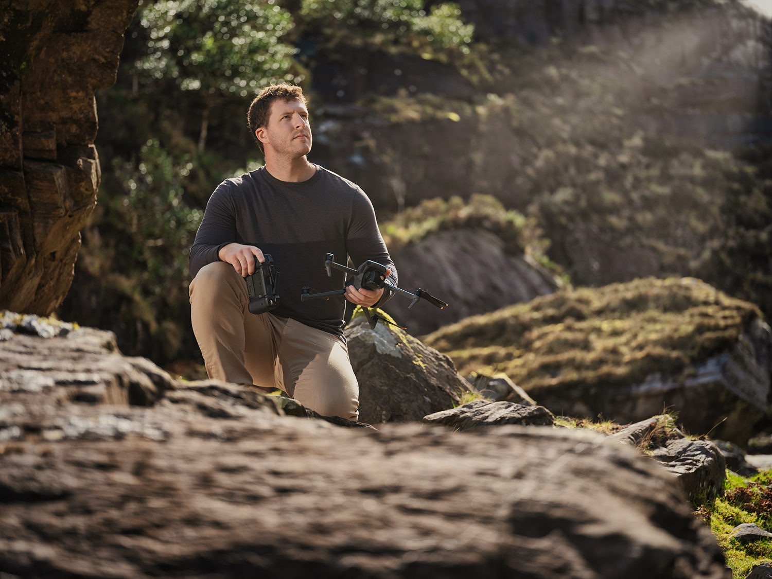 Shane Yeager, Ceo CineSalon crouches on rocks, holding equipment, looking up at foliage. Outdoor setting with sunlight.