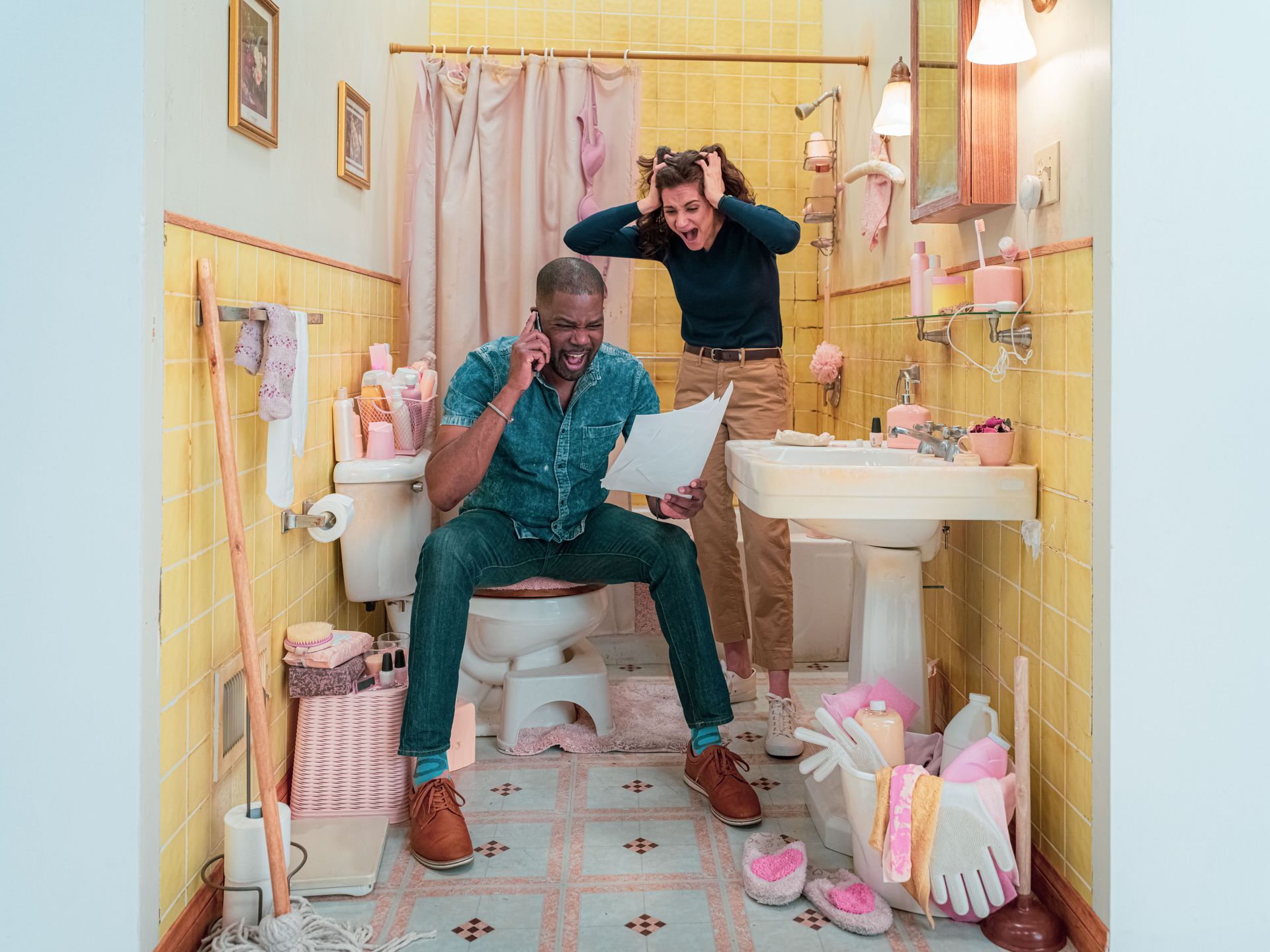 A man is sitting on a toilet in a bathroom while a woman stands behind him.
