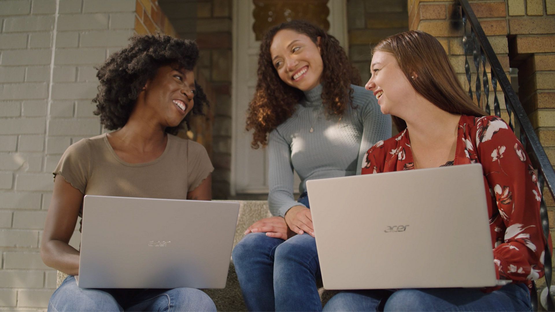 Three women are sitting on the steps of a house using laptops.