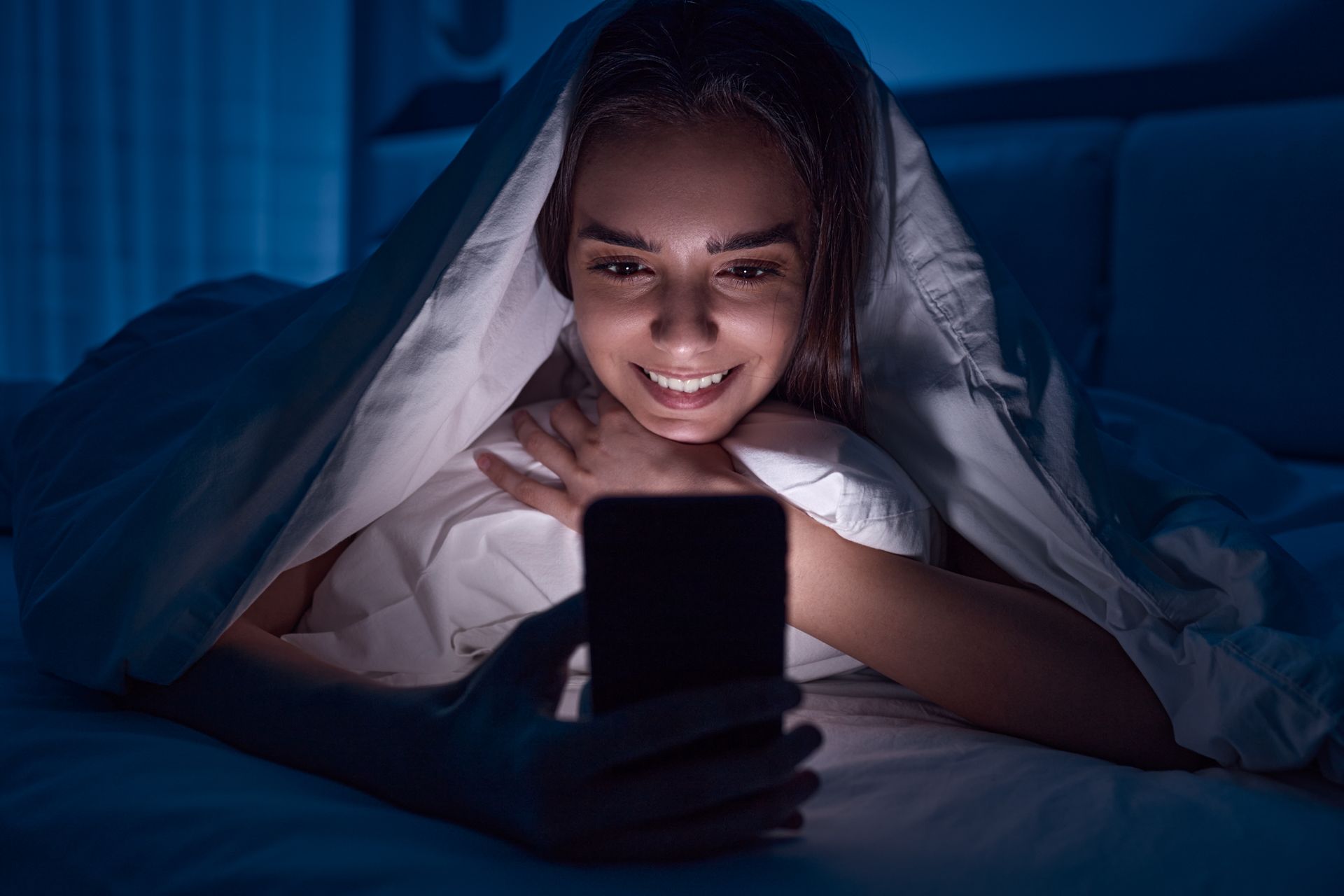 A woman is laying in bed under a blanket looking at her cell phone.