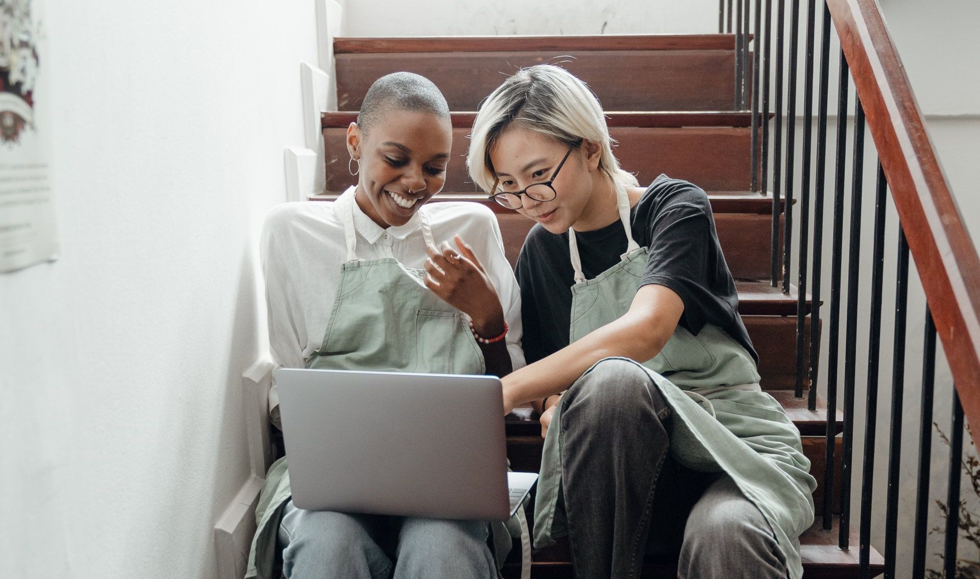 Two women are sitting on a set of stairs looking at a laptop.