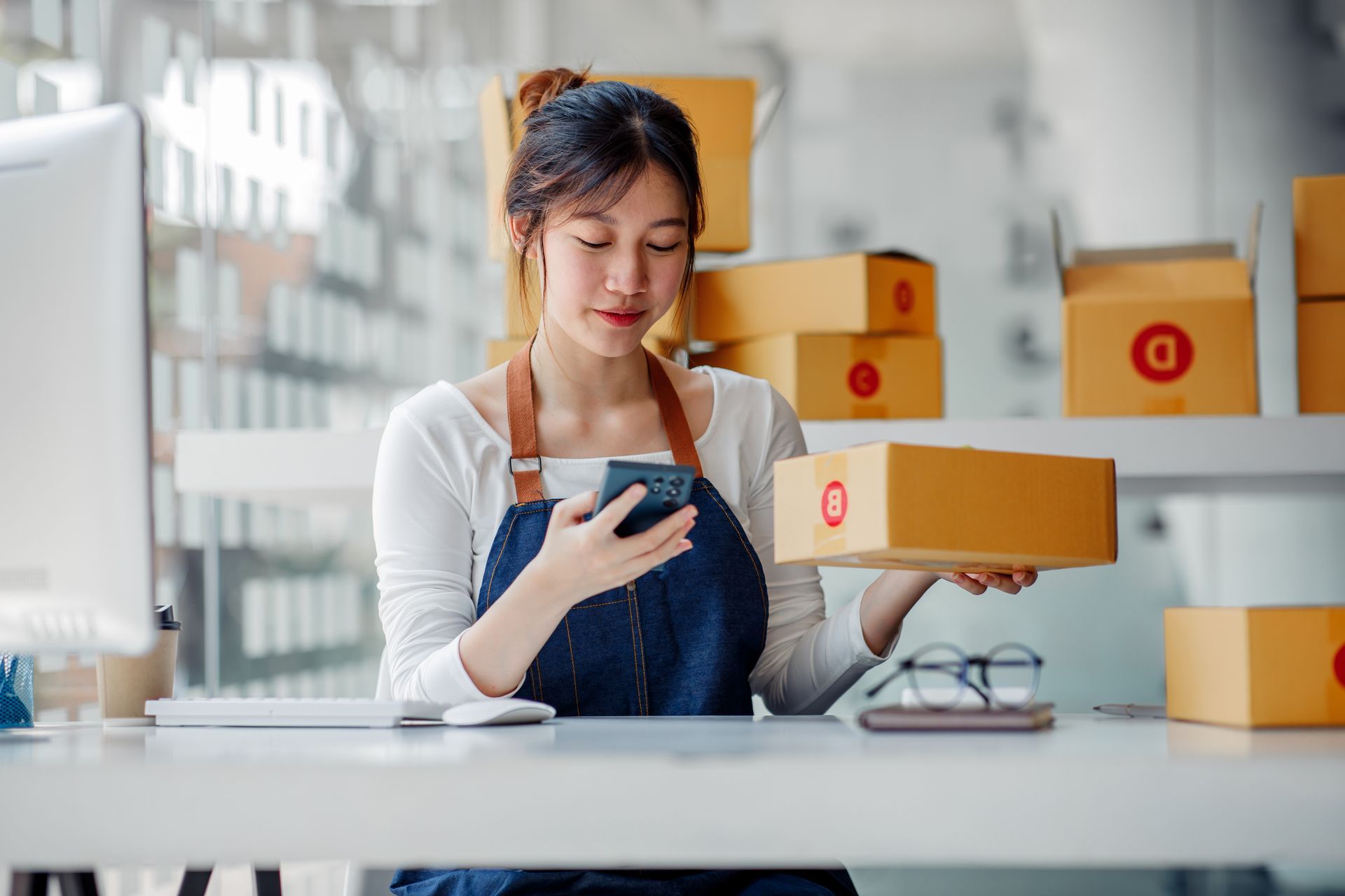 A woman is sitting at a desk holding a box and looking at her phone.