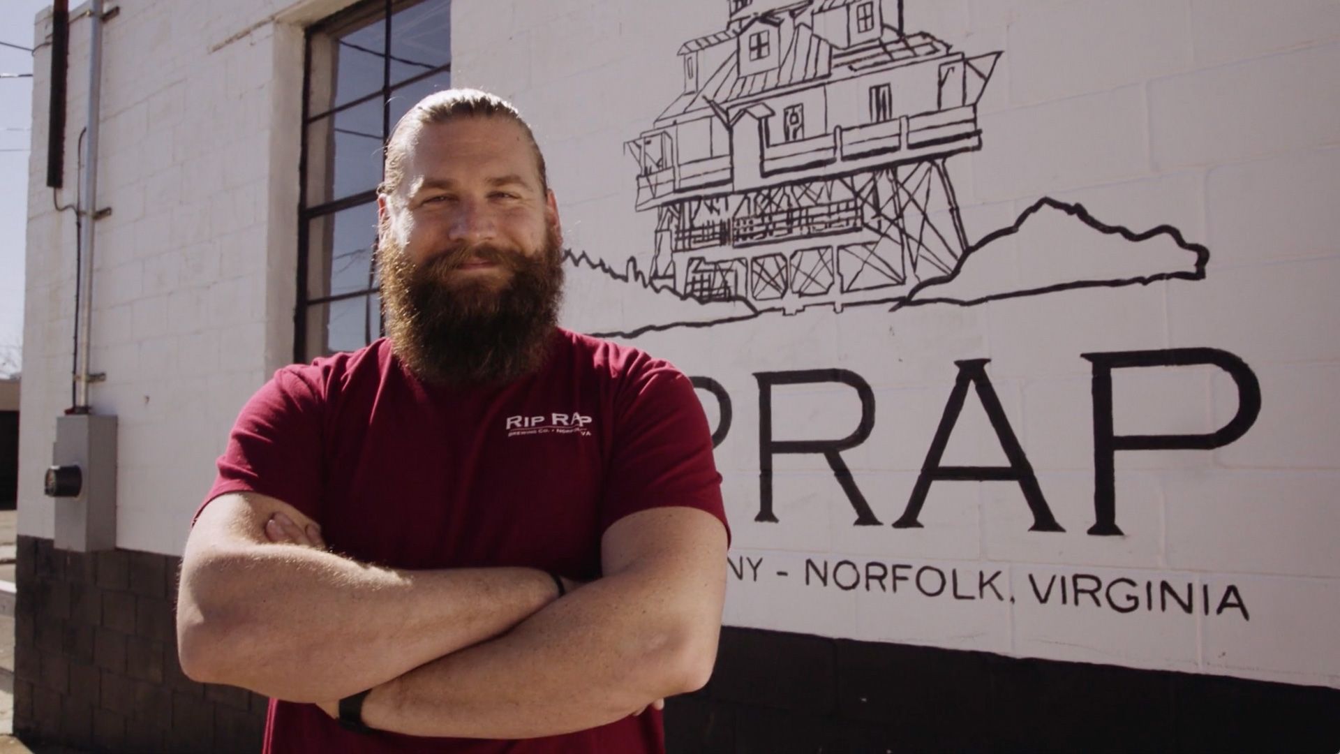 A man with a beard is standing in front of a building with his arms crossed.