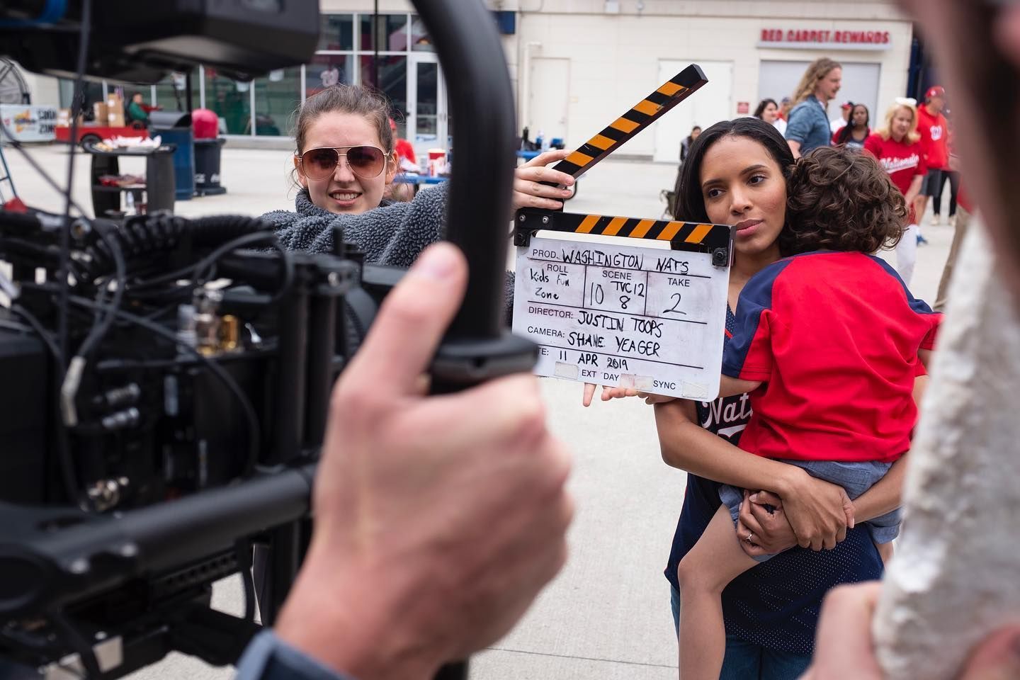 On-set scene: woman holds child and clapperboard; another operates camera, filming outdoors.