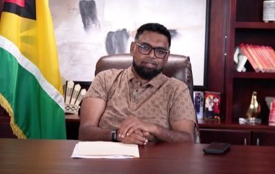 Man with glasses sits at a desk, Guyanese flag in the background.