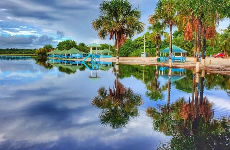 Lush waters reflect palm trees and turquoise gazebos under a cloudy sky.