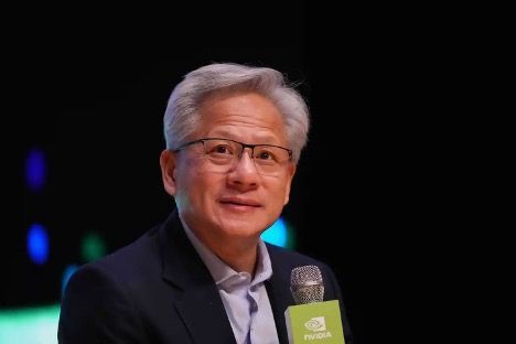 Jensen Huang, CEO of Nvidia, smiling, speaking into a microphone with Nvidia logo, dark background.