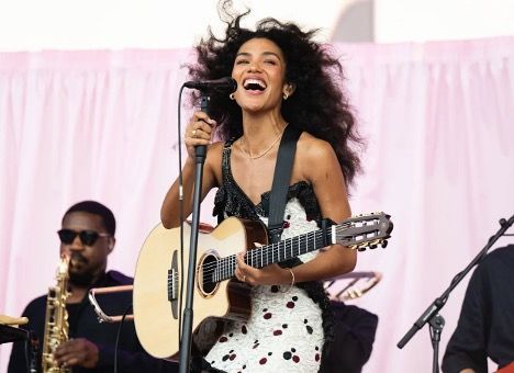 Woman singing and playing acoustic guitar onstage, smiling. Pink backdrop, band members visible.