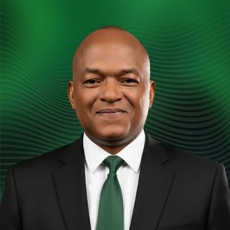 Man in suit, green tie, smiling, against a green, patterned background.