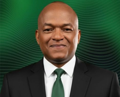 Man in black suit and green tie smiles in front of a green background.