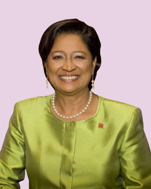 Woman in a green suit and pearl necklace smiles, set against a pink background.