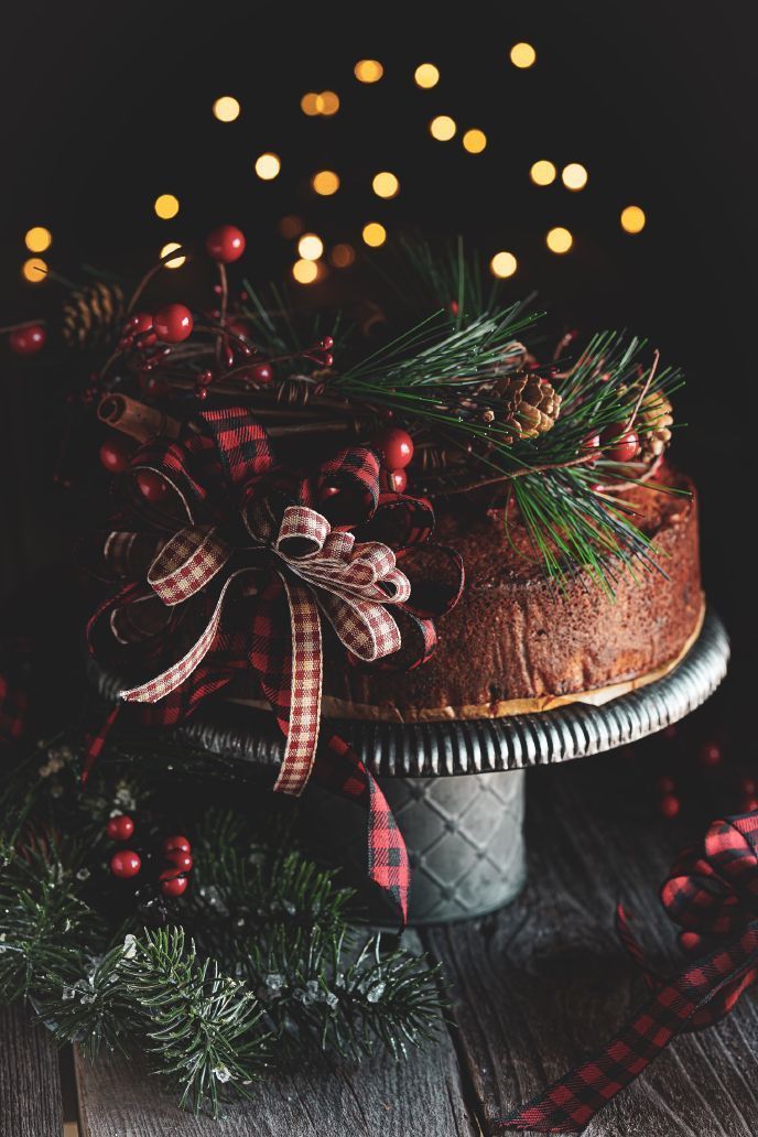 Christmas cake on a pedestal, decorated with pine, berries, and a plaid bow, with blurred lights in the background.