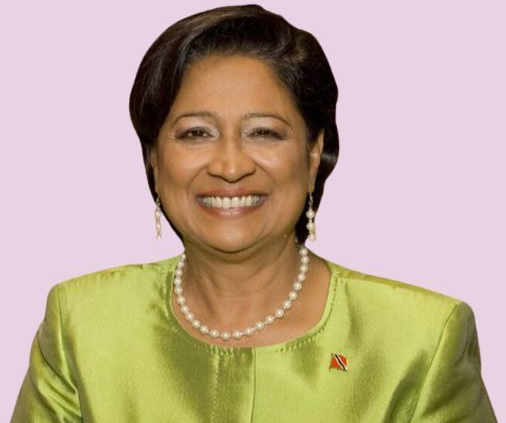 Kamala Persad-Bissessar smiles, wearing a green jacket and pearl necklace, against a purple background.
