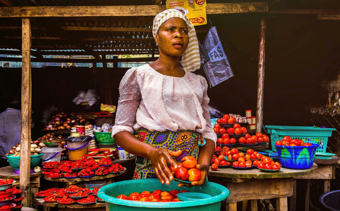 Woman at a market stall selling tomatoes, sorting them into a bowl. Tomatoes are red.