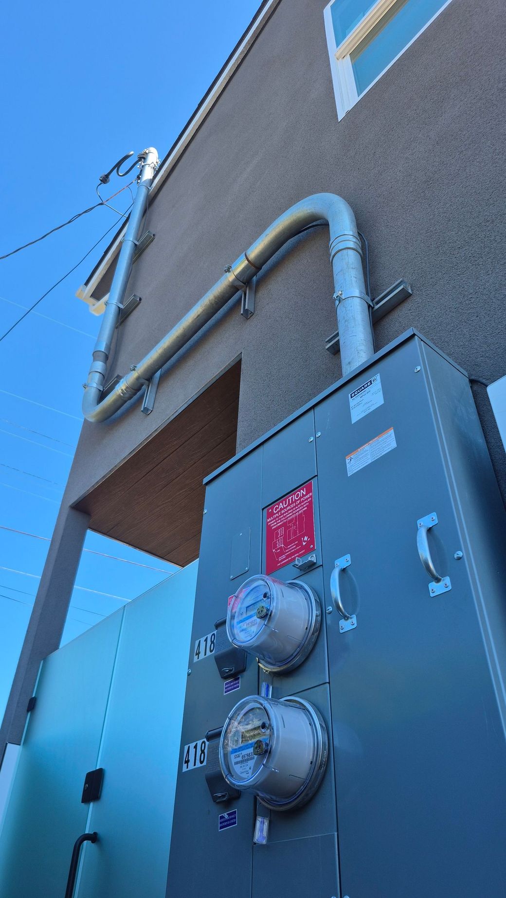 Outdoor utility meter panel on a stucco building wall with gray pipes and a blue sky above