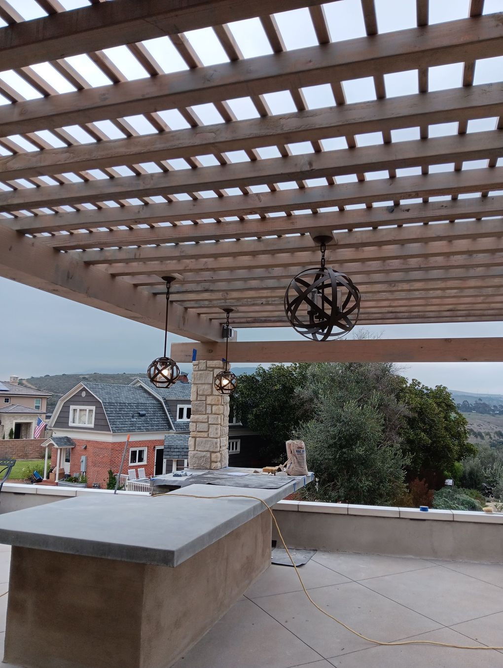 Covered patio with wooden pergola, stone counter, hanging lights, and hillside view beyond.