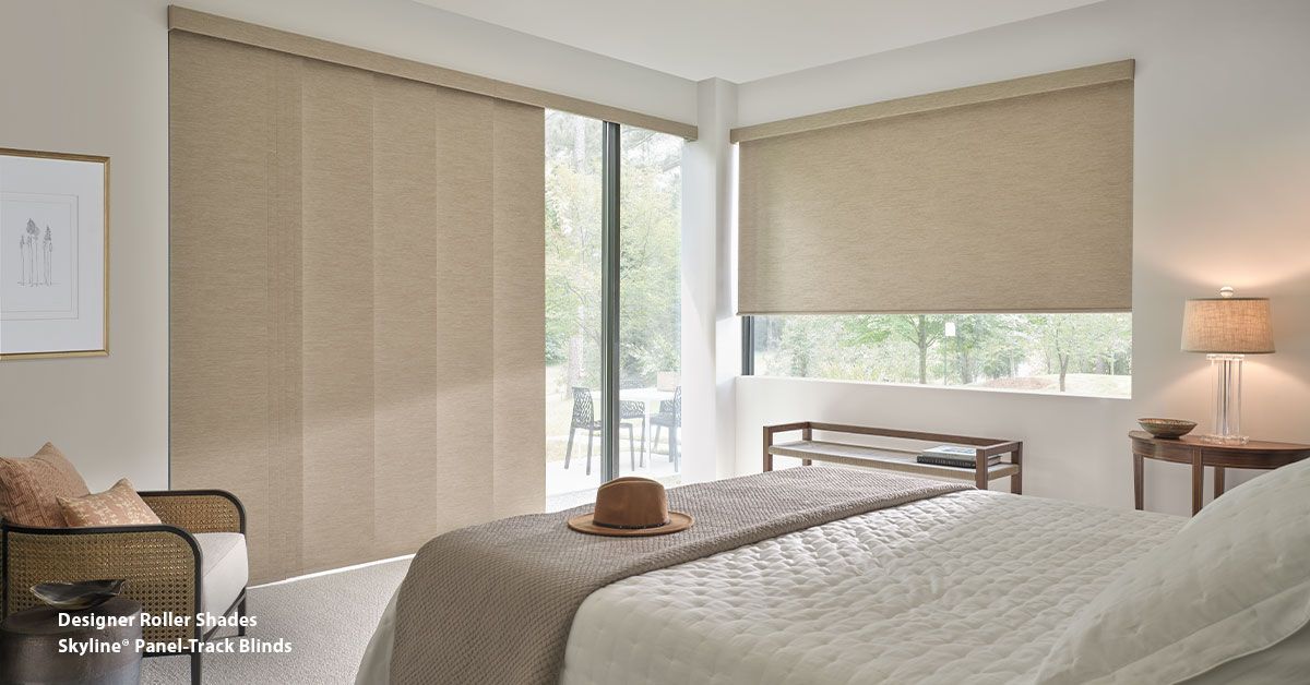 Bedroom with bed, panel blinds, and a roller shade. A lamp sits by a table.