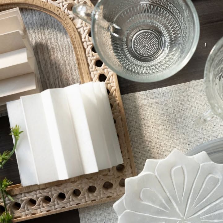 Overhead view: white folded square, clear teacup, patterned coasters on a woven tray, light neutral tabletop.