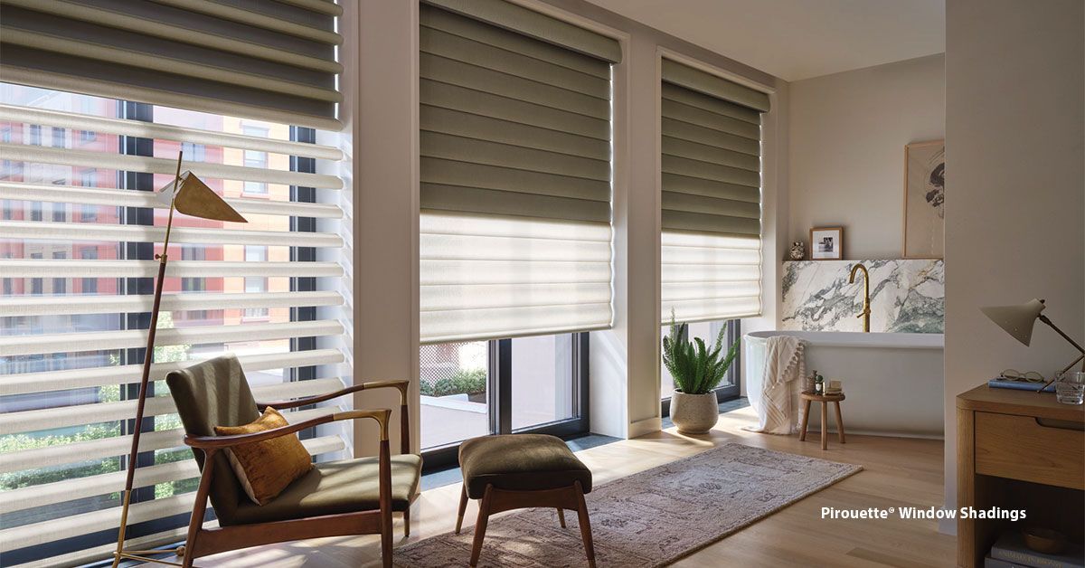 Room with three windows, each with shades. A chair, ottoman, lamp, and rug are in the room. Natural light.