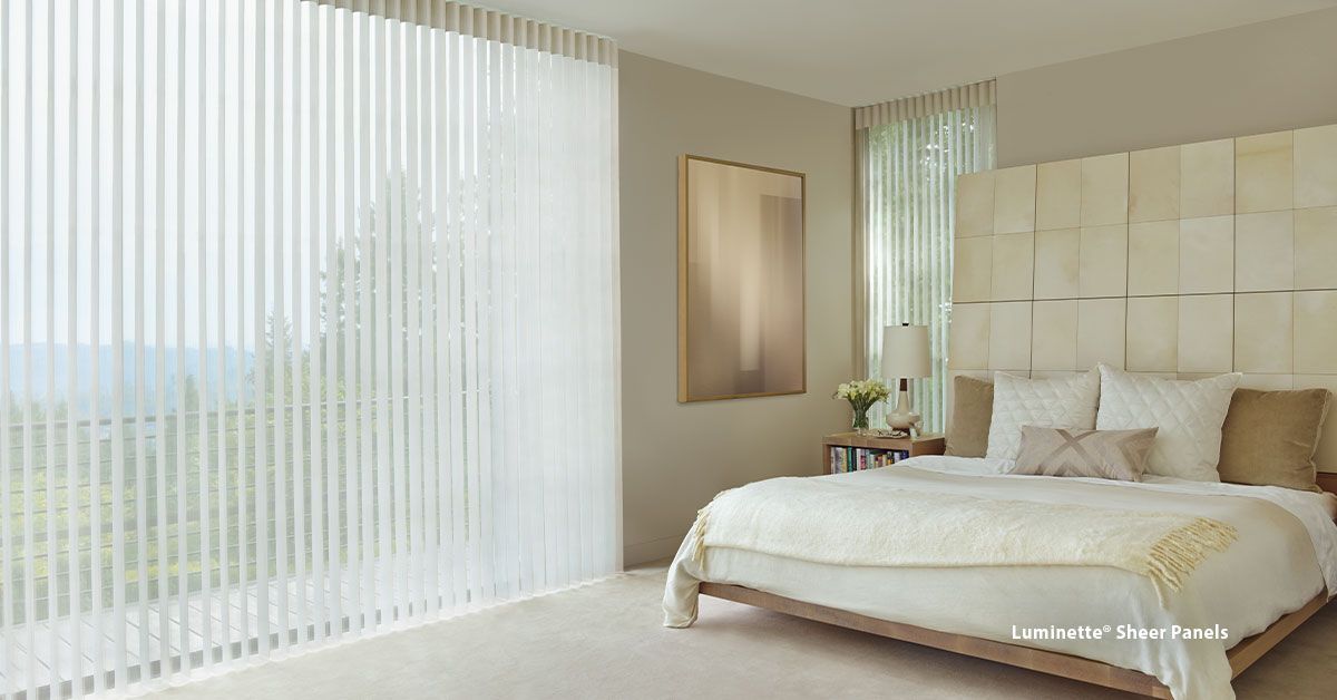 Bedroom with large window, vertical blinds, and a bed with a large headboard, all in neutral tones.