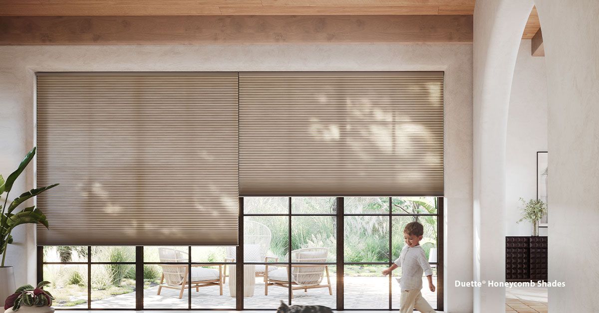 Child walks past windows with neutral-toned blinds, bright sunlight, and outdoor furniture visible.