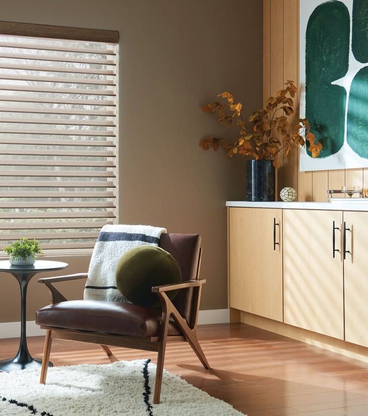 Cozy room with leather chair, window blinds, and abstract art. Warm brown walls, wood flooring, and a round pillow.