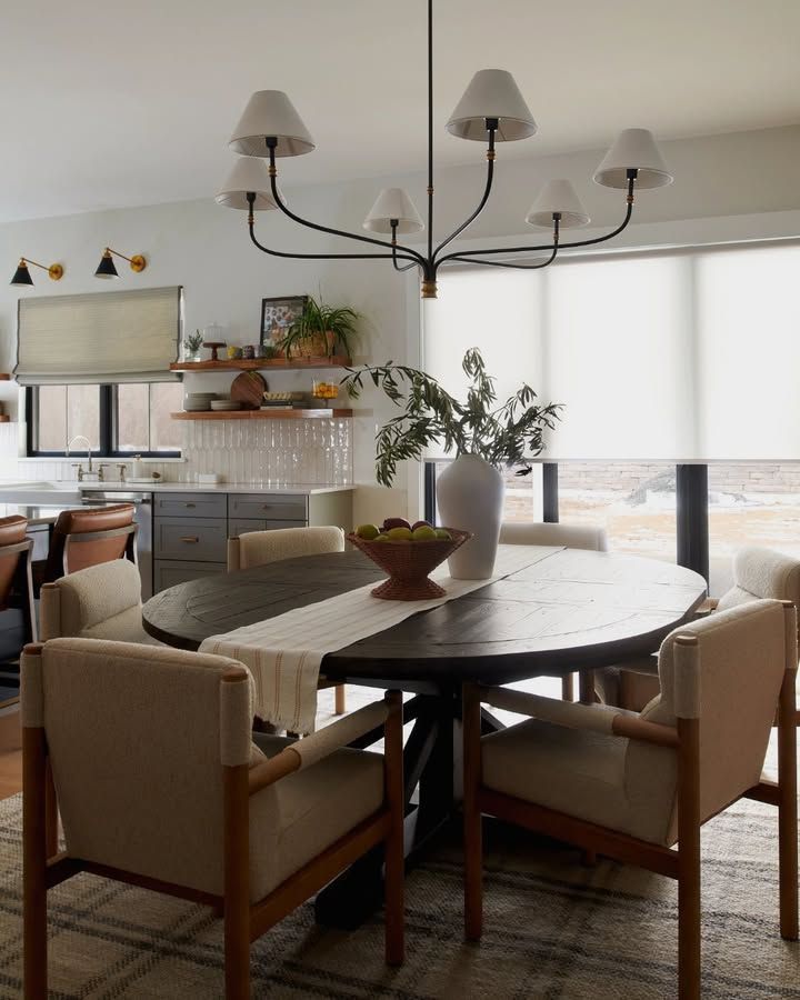 Dining room with oval table, chairs, and chandelier; neutral tones; plants and window coverings present.