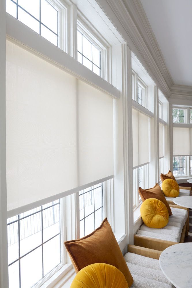 Cream-colored roller shades cover the windows of a bright, white-framed window seat with throw pillows.